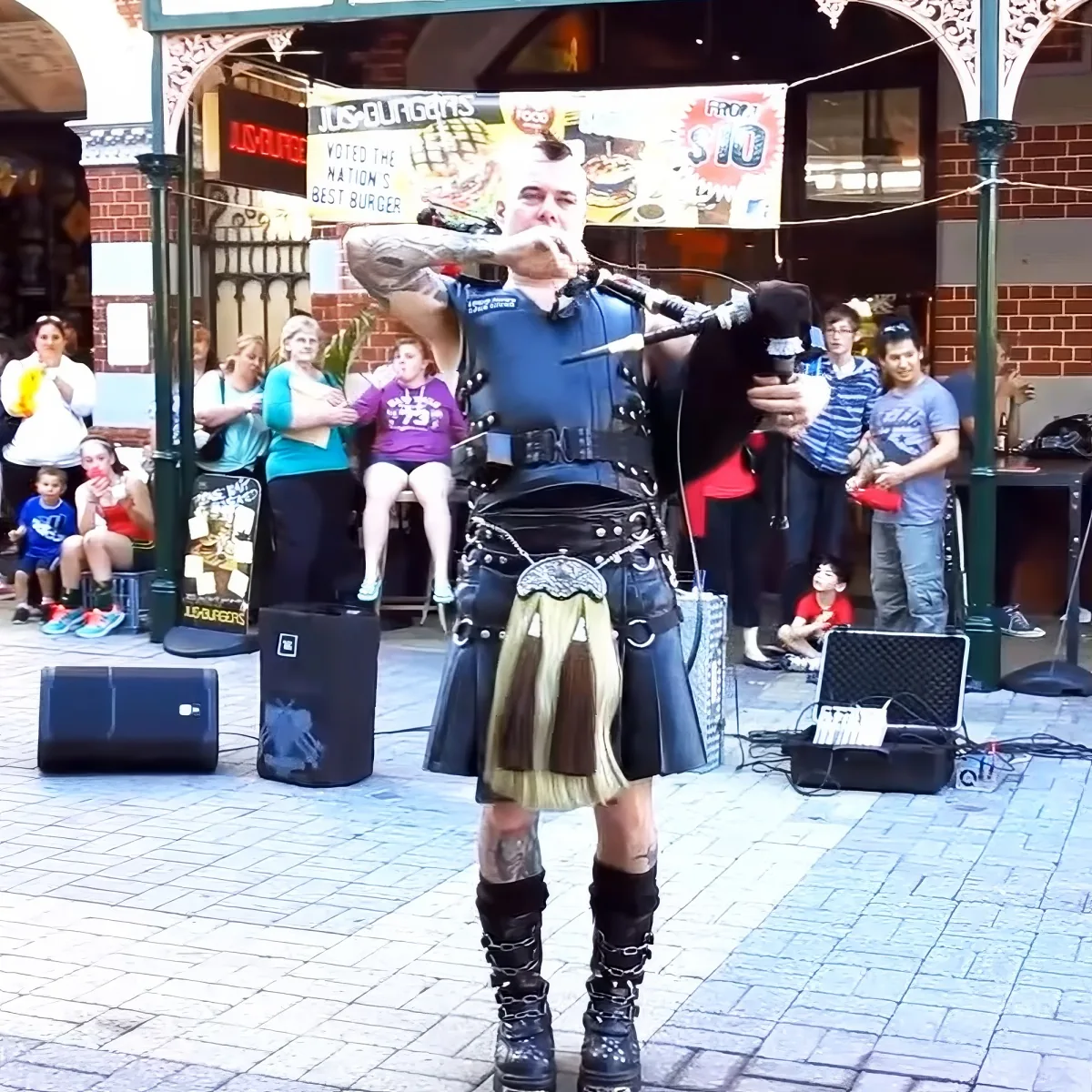 A man in a kilt plays bagpipes on a street stage, surrounded by spectators and a burger sign.