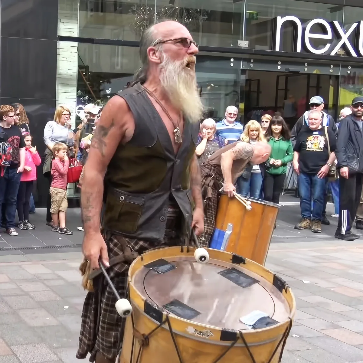 A man with a long white beard and tattoos plays a large drum on a city street, surrounded by an audience.