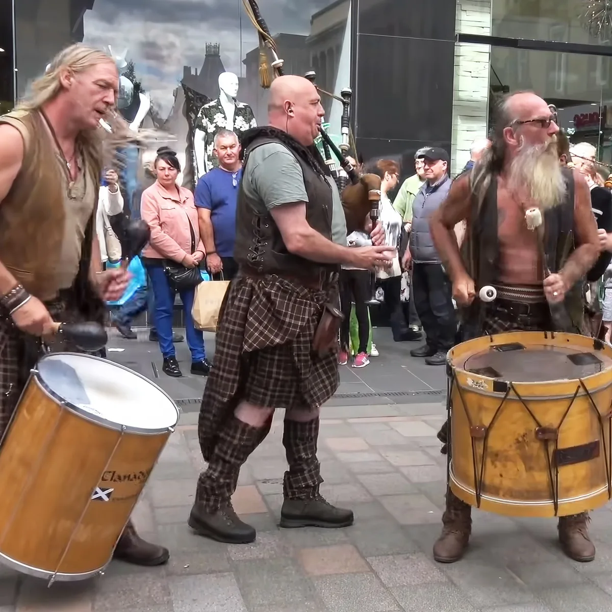 Three men in kilts play drums and bagpipes on a street, surrounded by onlookers.