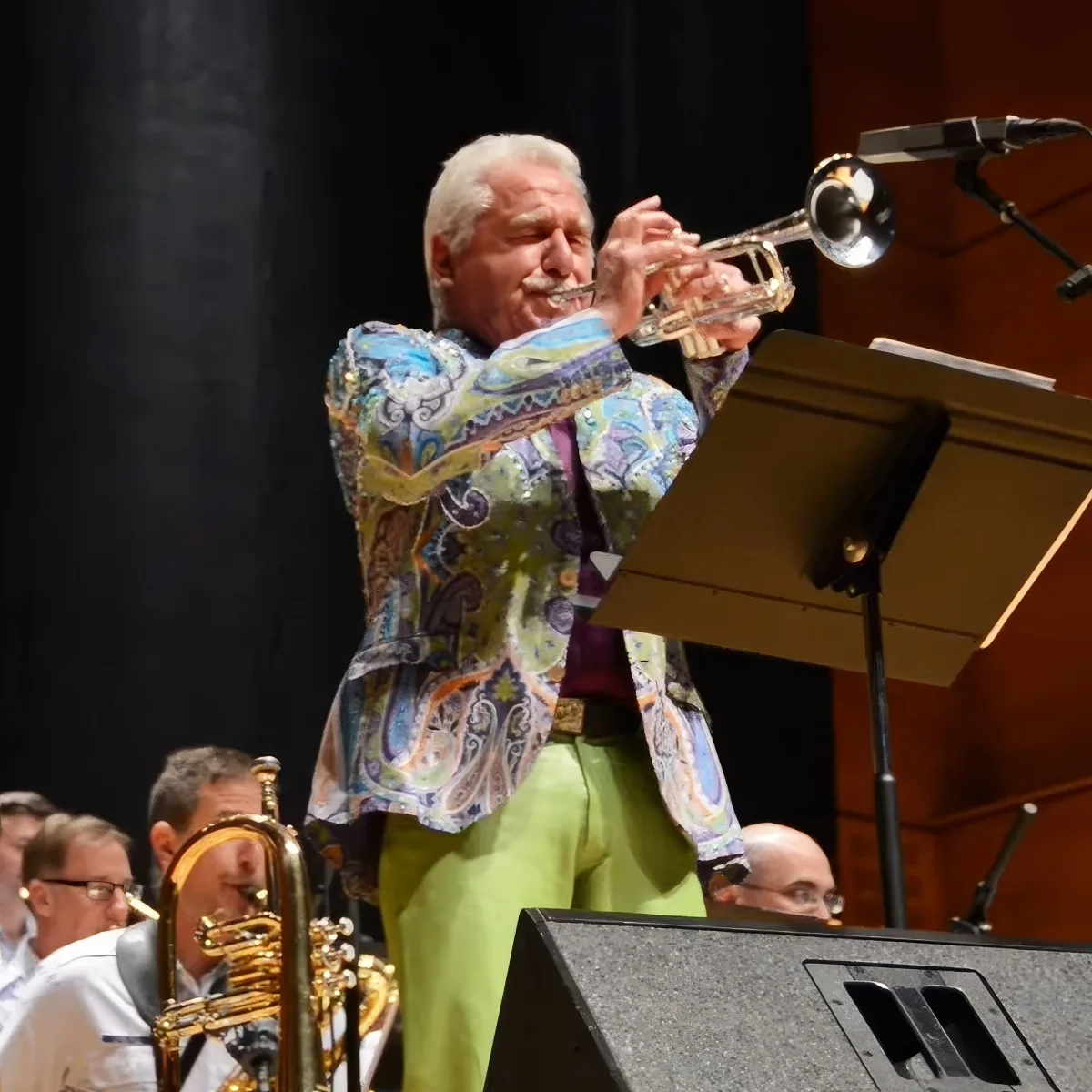 A man in a colorful paisley jacket plays a trumpet on stage, surrounded by other musicians.