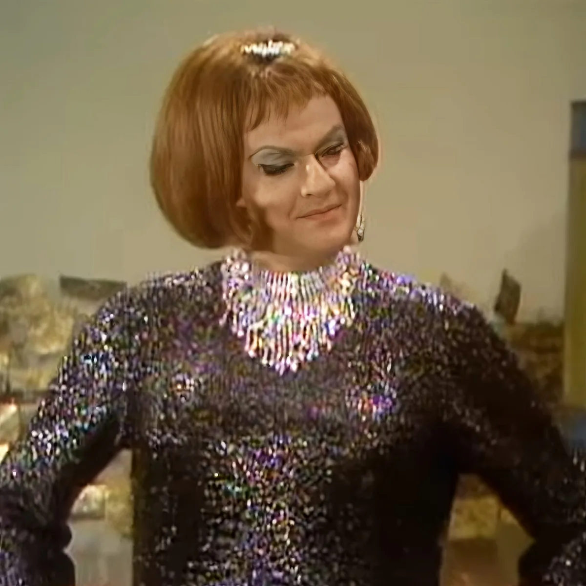 A person with short, styled hair and a sequined outfit is looking down.