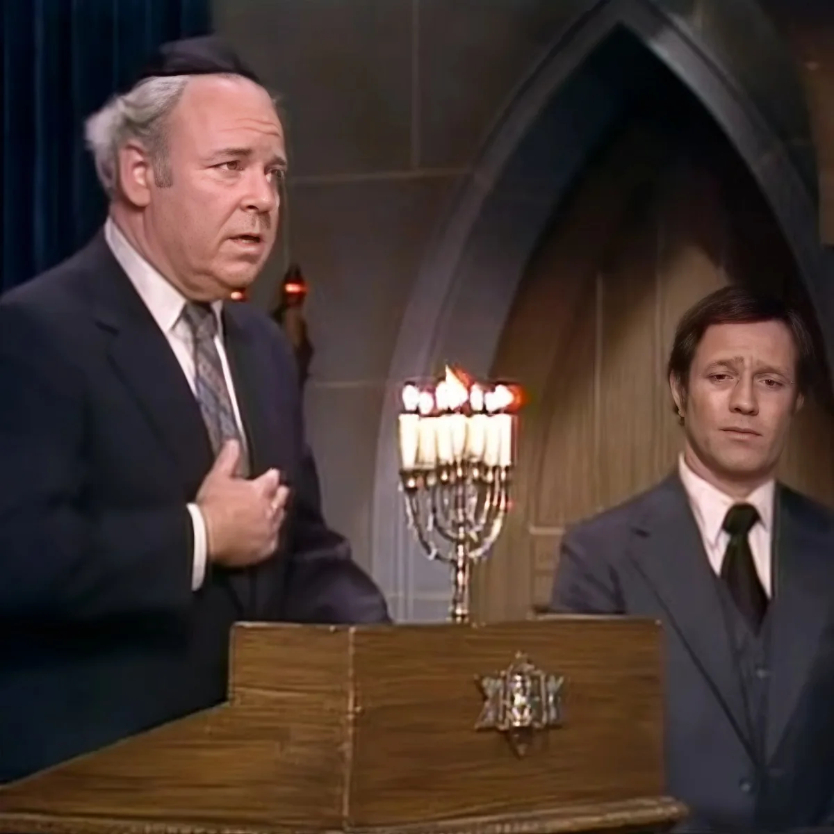 A man in a suit gestures beside a menorah, another man in a suit stands nearby, both in a formal setting.