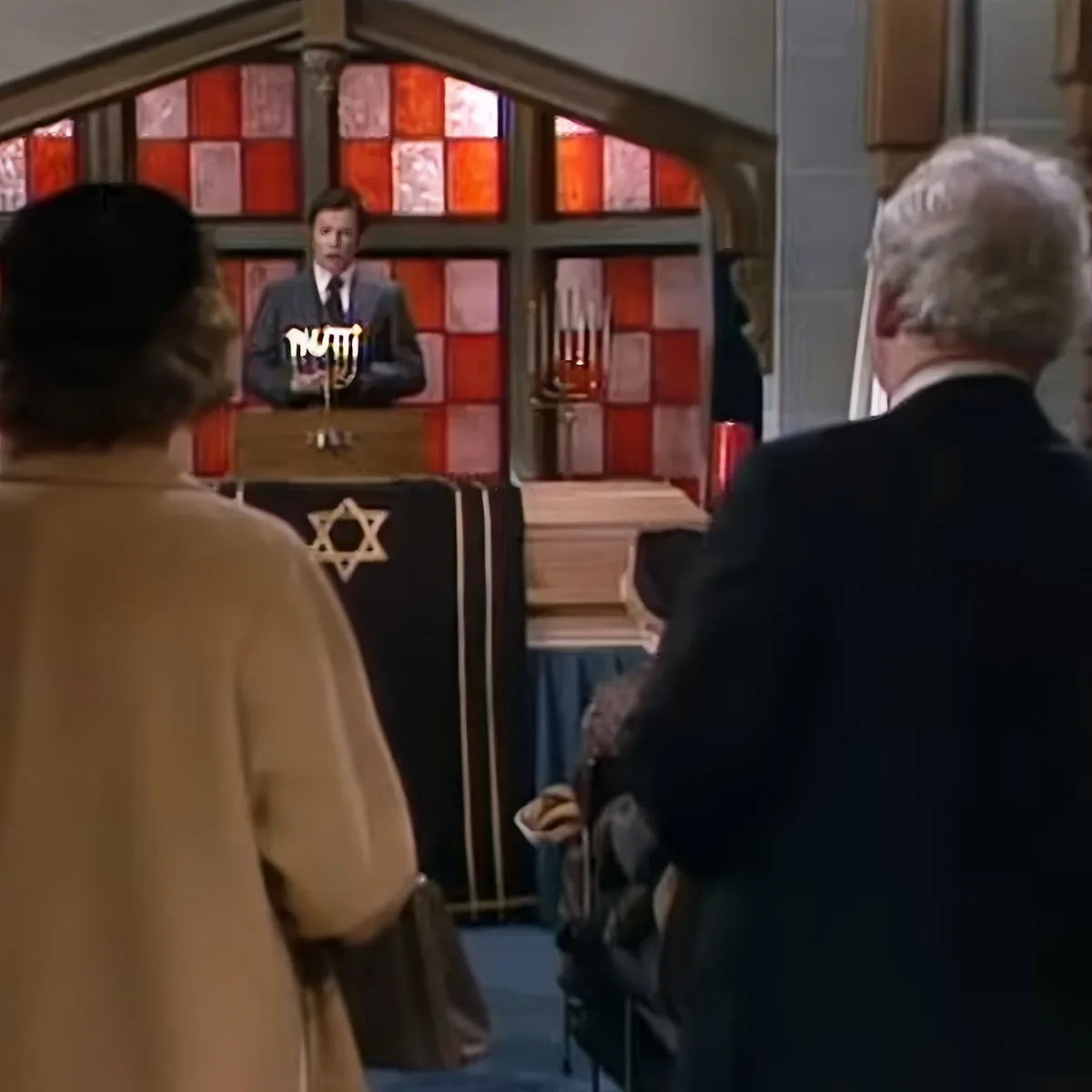 A man in a suit stands at a podium in a room with stained glass, addressing a seated audience.