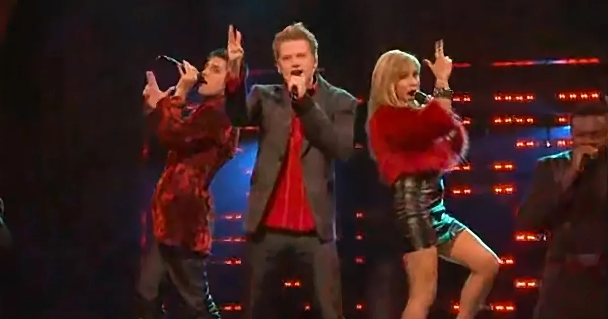 A man in a suit sings into a microphone, flanked by two women in red and black outfits, performing on stage.