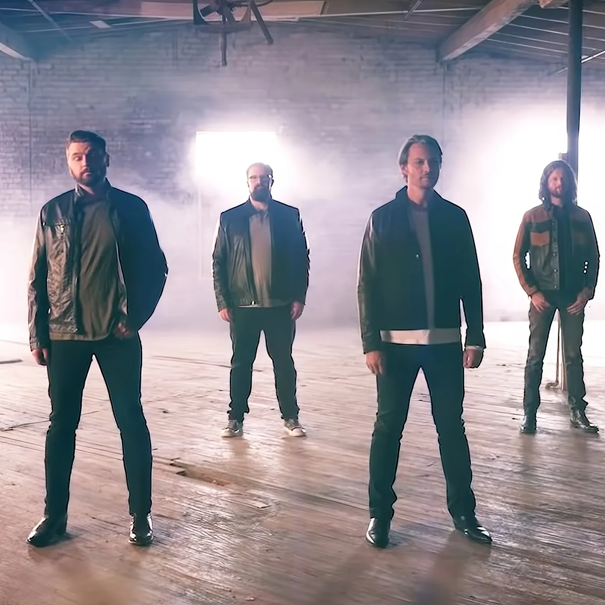Four men in leather jackets stand in a dimly lit, smoky warehouse setting.
