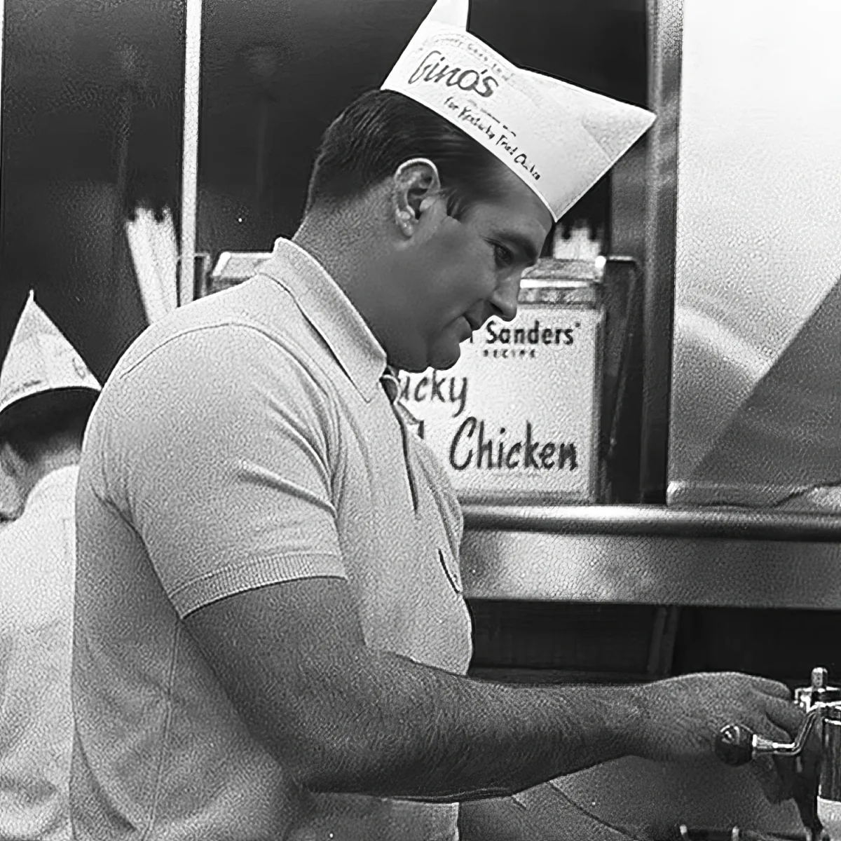 A man wearing a paper hat and a polo shirt is working in a kitchen.