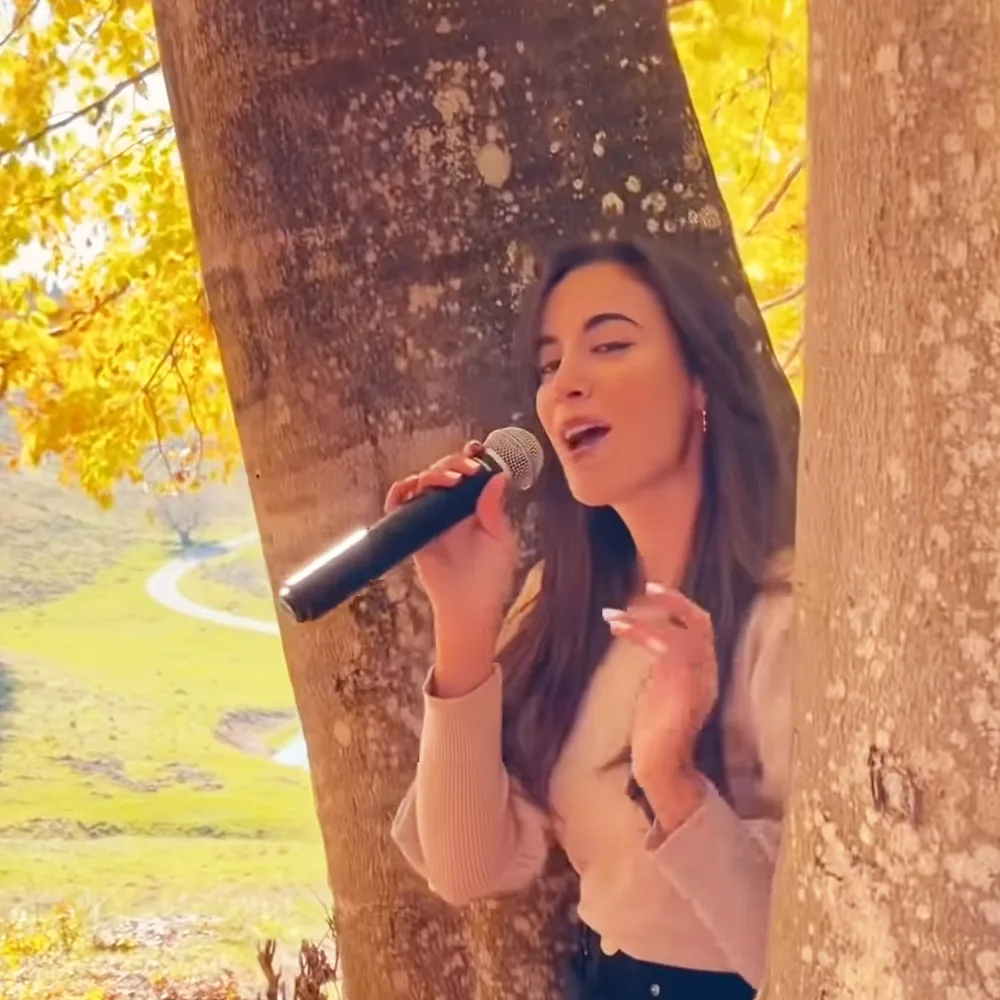 A person sings into a microphone, surrounded by autumn foliage and tree trunks.