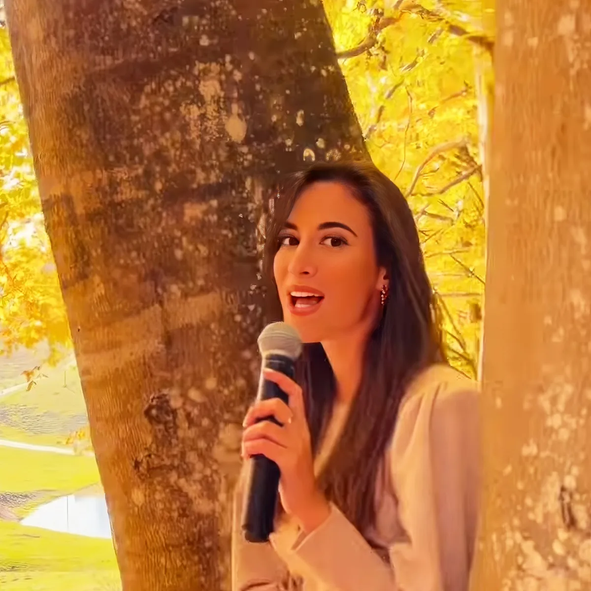 A woman with long dark hair holds a microphone, standing outdoors with trees and golden light in the background.