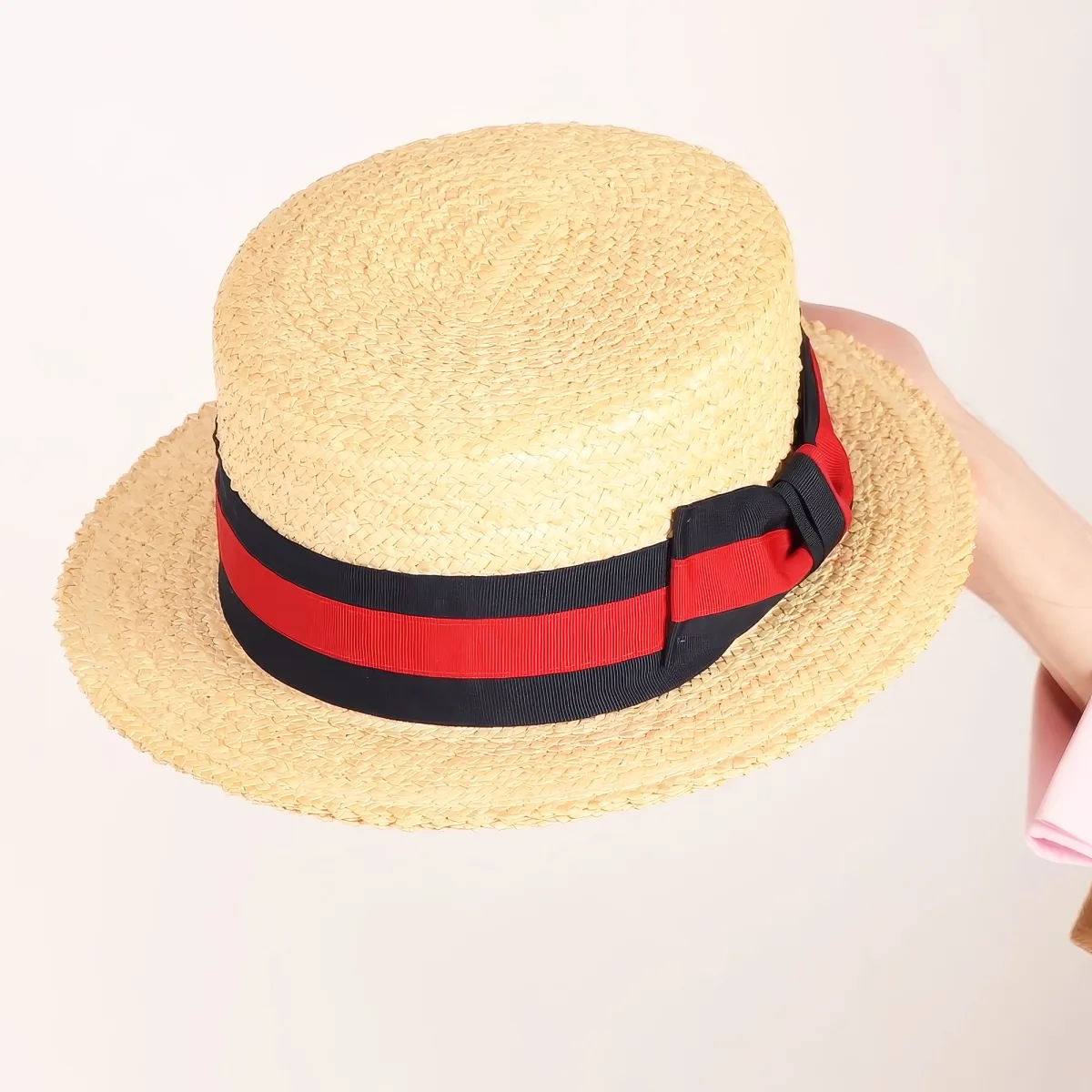 A straw hat with a black and red ribbon around its base is being held by a person in a pink sleeve.