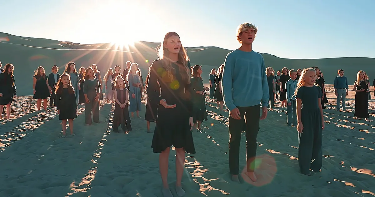 A young woman in a black dress stands in front of a group of people singing on a sandy beach under a bright sky.