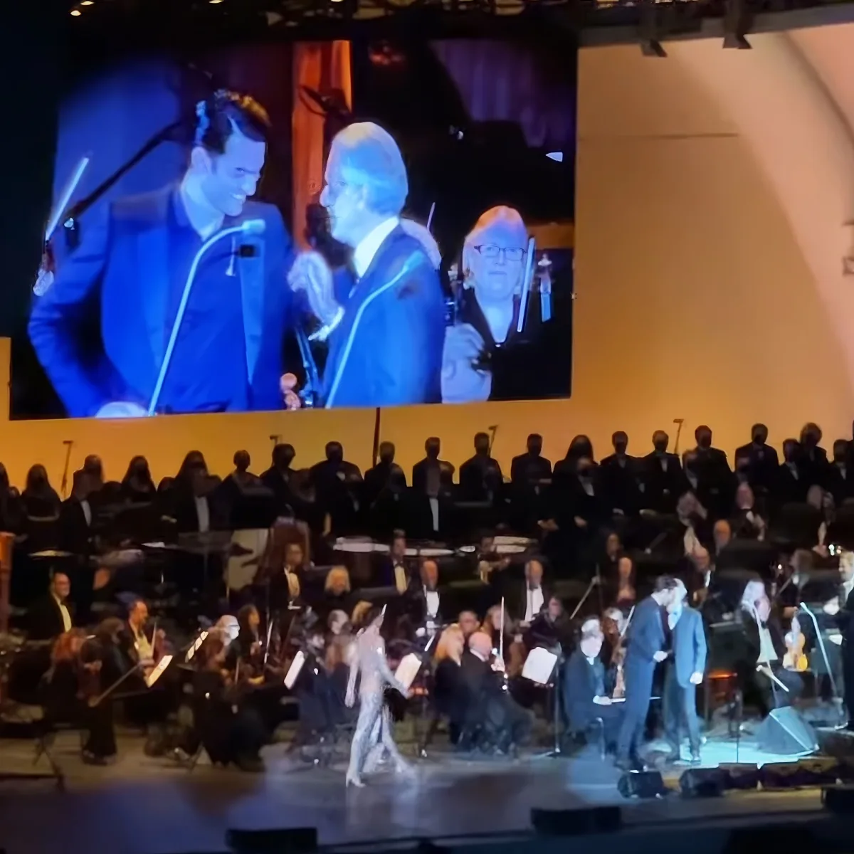 A conductor leads an orchestra on stage, with a large screen displaying two men in blue suits.