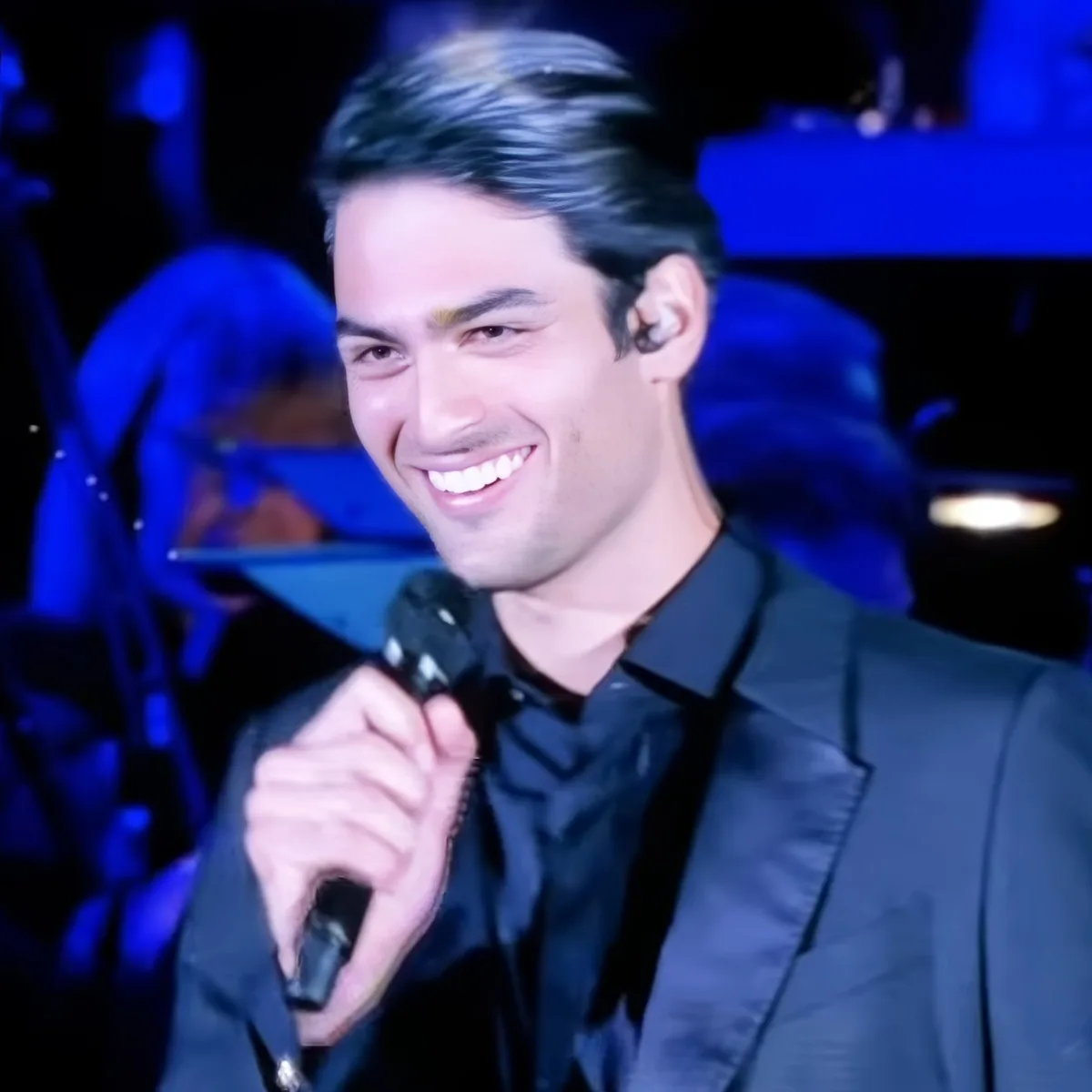 A man in a black suit smiles while holding a microphone on stage.