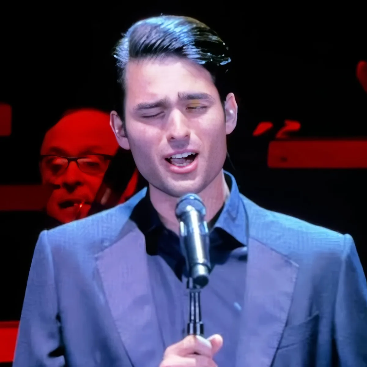 A man in a suit sings into a microphone, with a red-lit figure in the background.