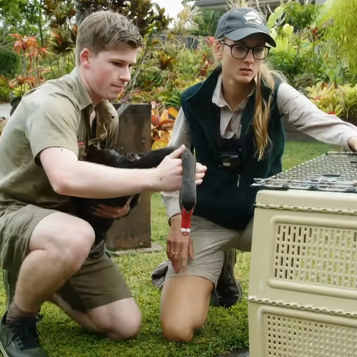 A man and woman in uniform hold a small animal, possibly a bird, near a cage in a grassy area.