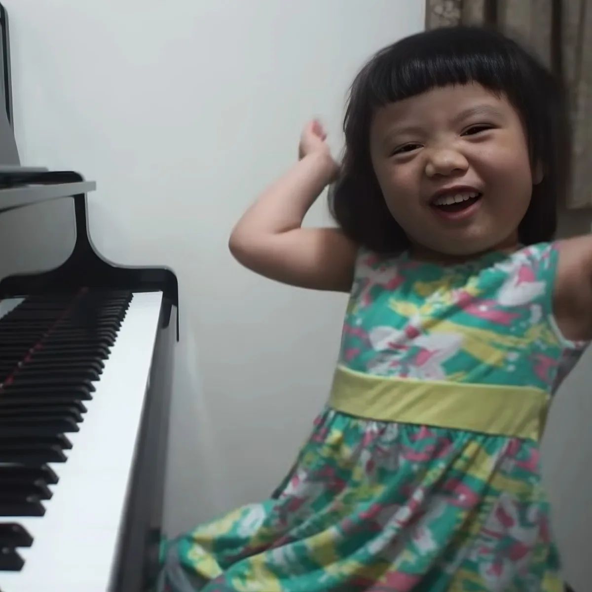 A young child in a colorful dress is smiling and pointing towards the camera, with a piano visible on the left.