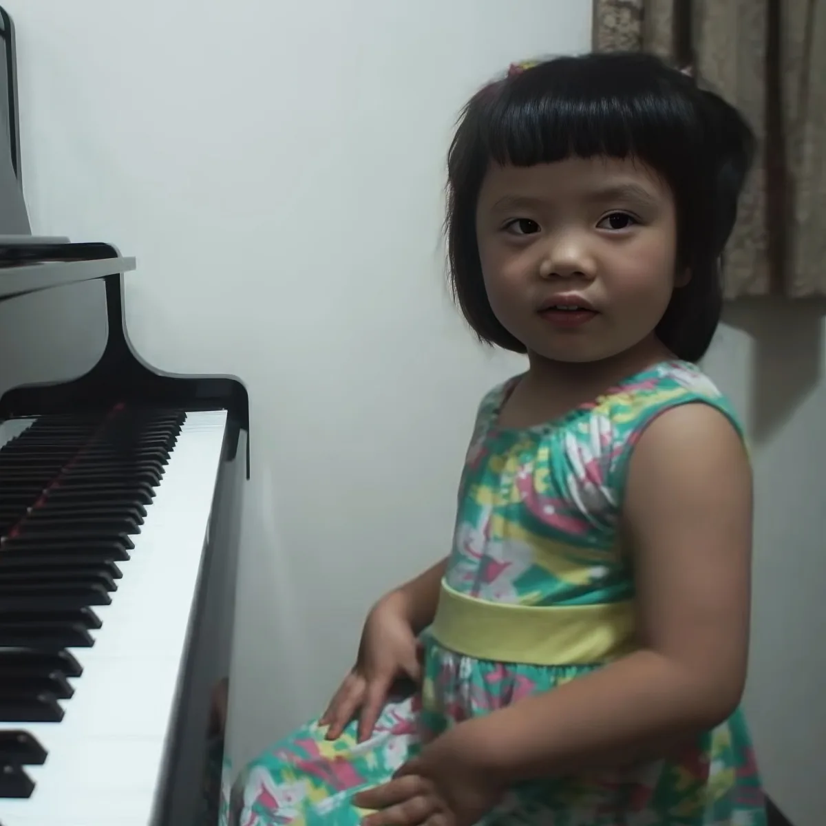 A young child in a colorful dress stands near a piano, looking slightly to the side.