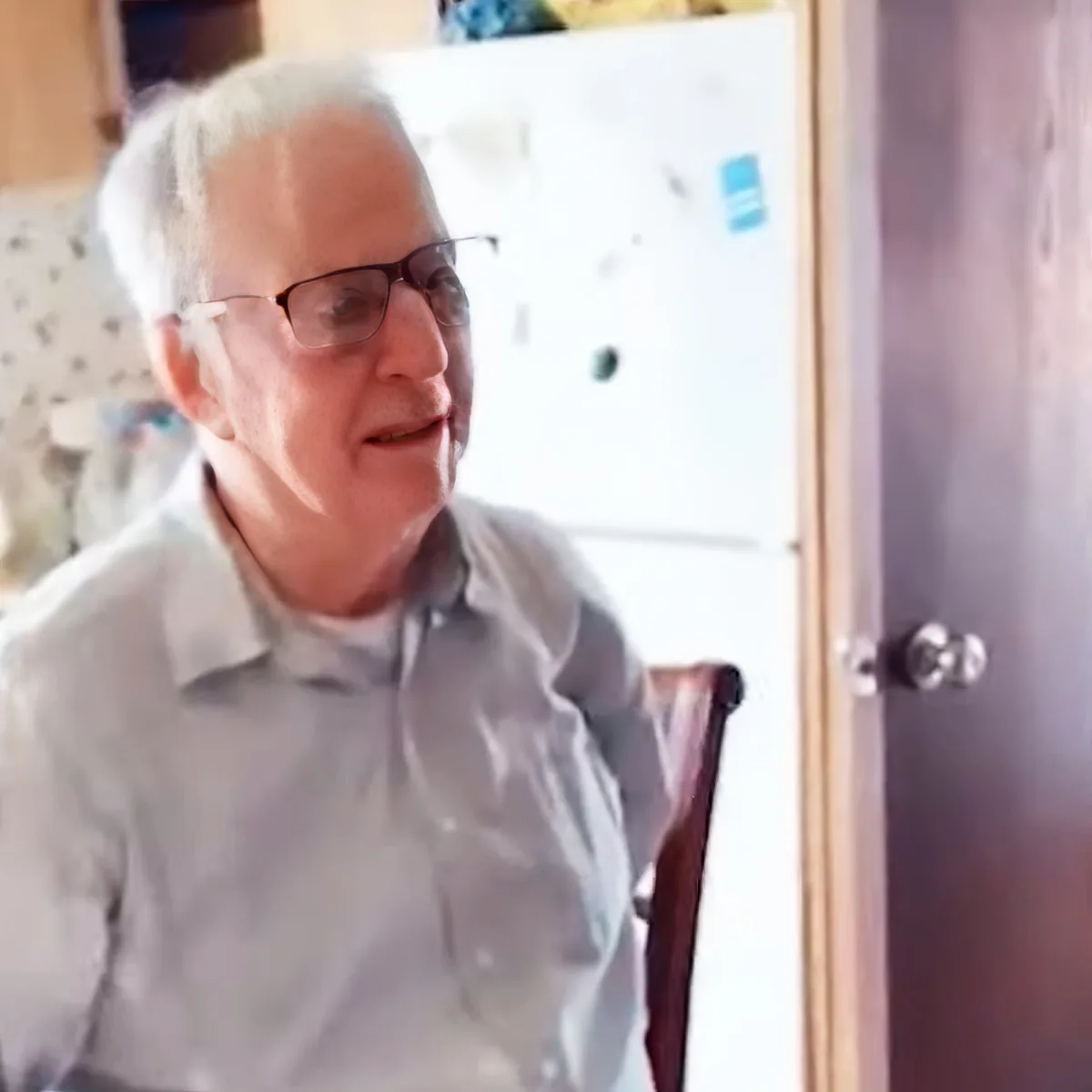 An older man wearing glasses and a light shirt sits in a chair, smiling, with a white refrigerator and door in the...