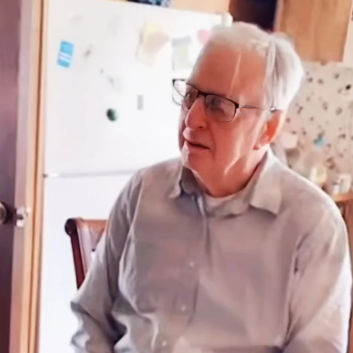 An older man wearing glasses and a light shirt sits in a chair, smiling, with a whiteboard and door in the background.