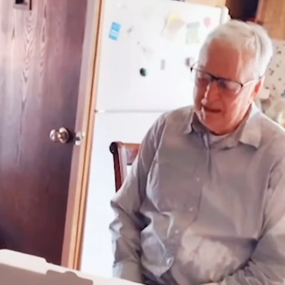 An older man with white hair and glasses sits at a table, looking down, in a room with a whiteboard and door in the...
