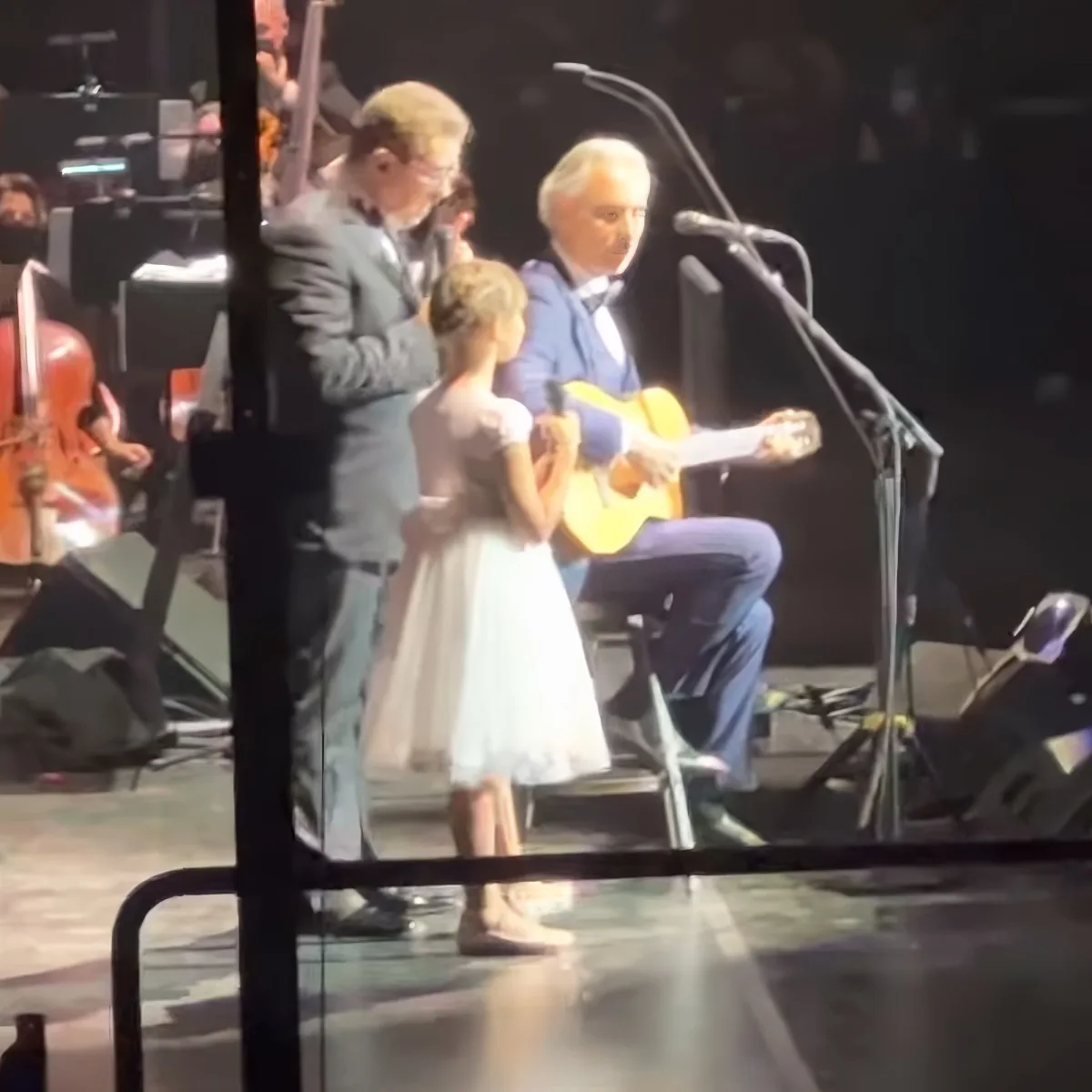 A man in a blue suit plays guitar on stage with a young girl in a white dress.