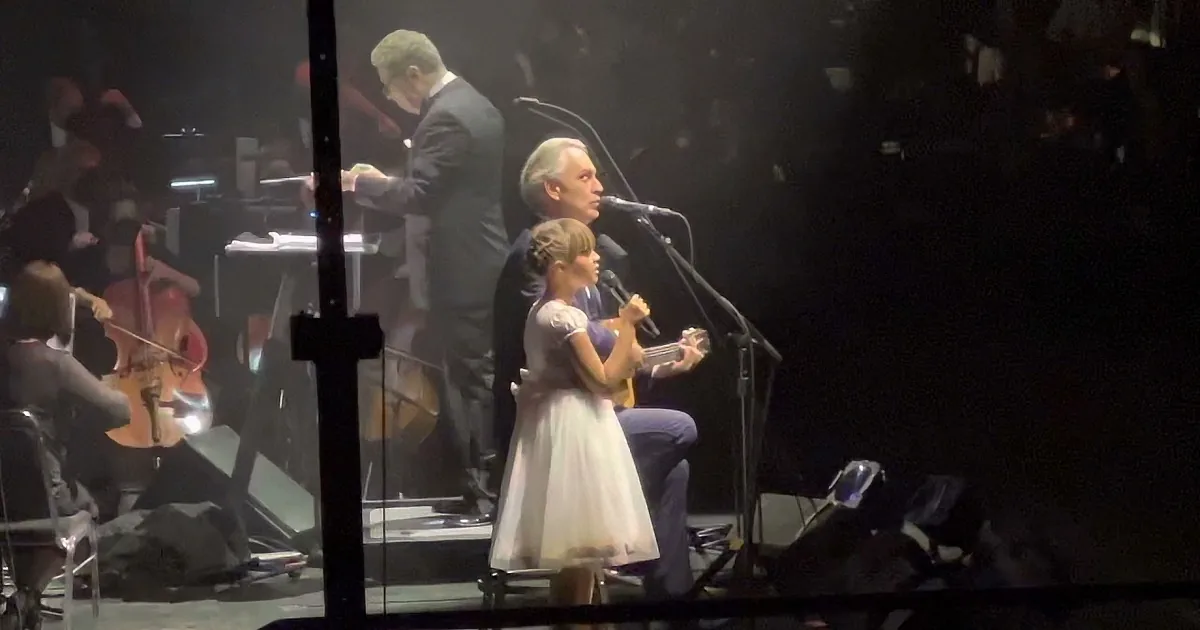 A young girl in a white dress sings into a microphone while an older man accompanies her on stage.