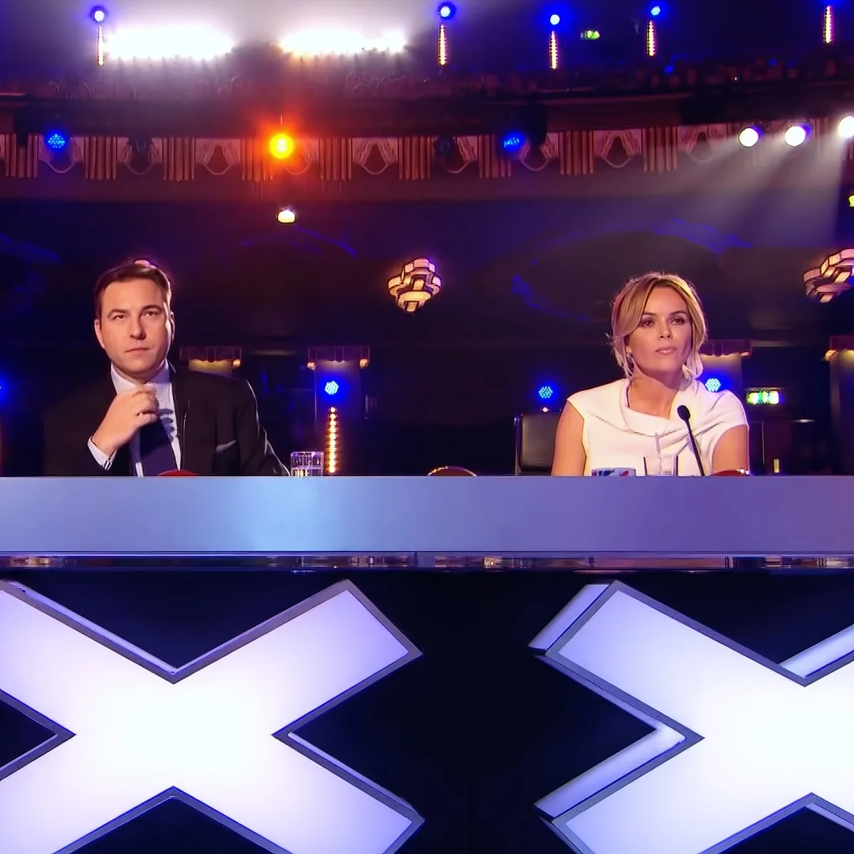 Two judges sit at a table with a large "X" illuminated in the foreground, likely from a talent show.