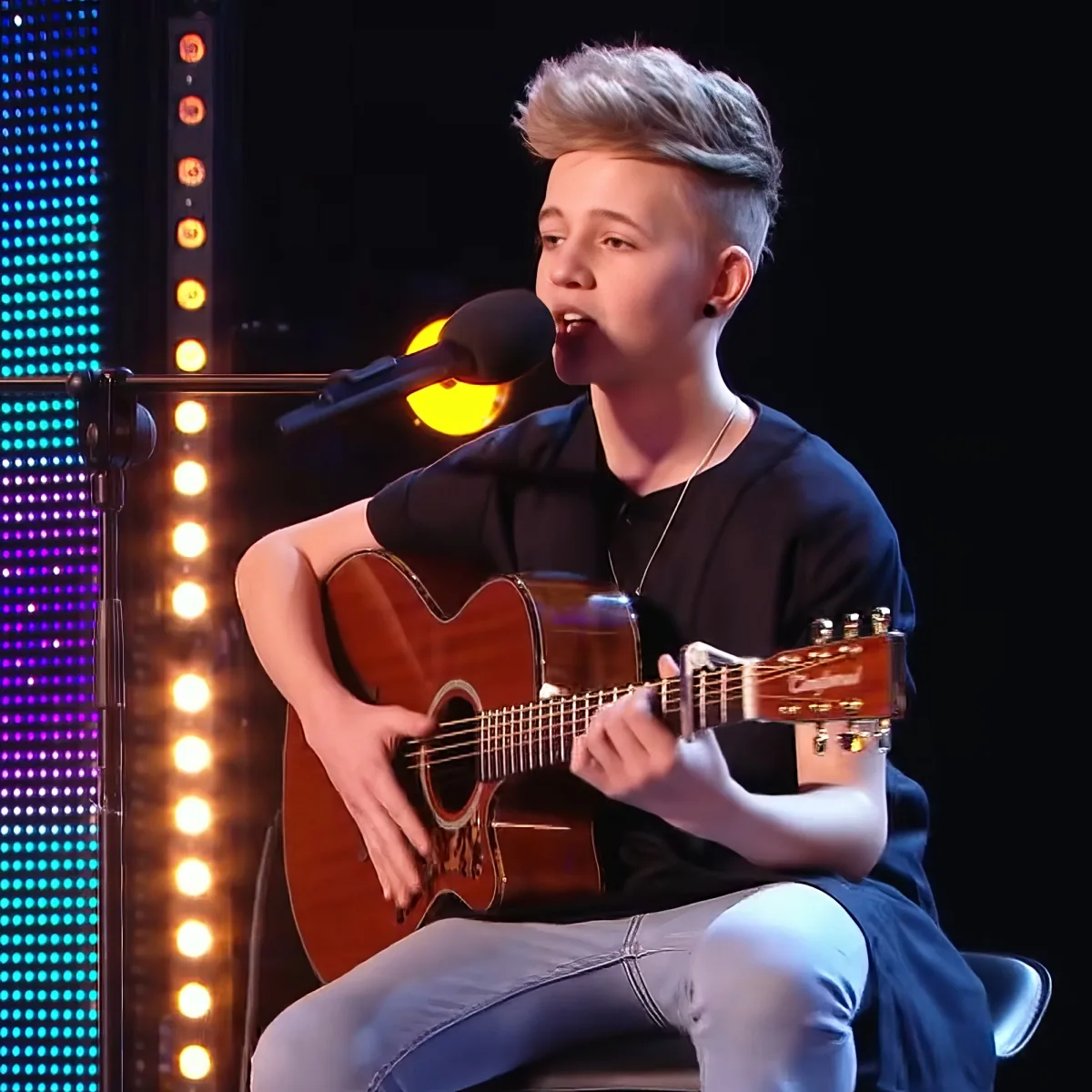 A young person plays an acoustic guitar and sings into a microphone on a stage with colorful lights.