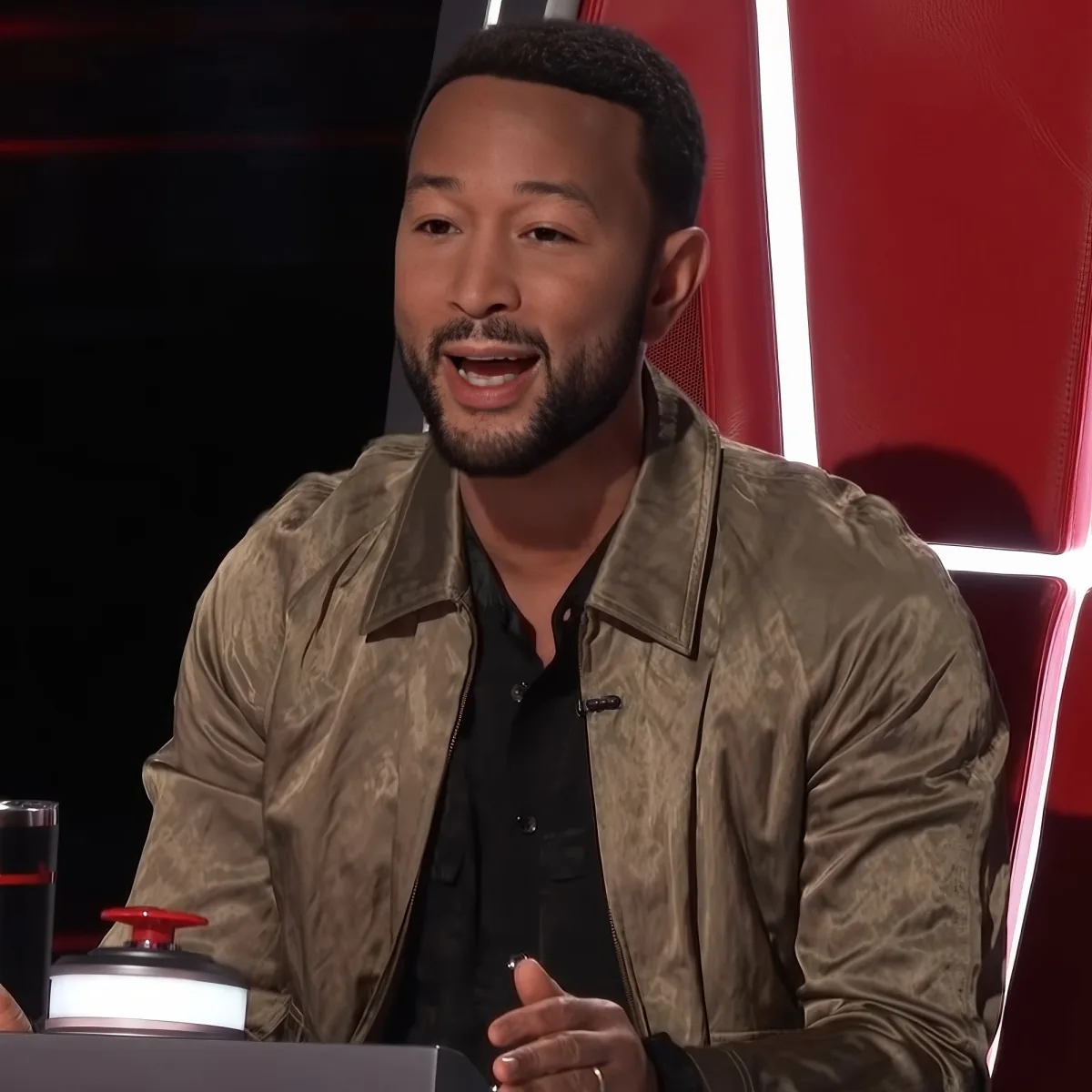 A man in a brown jacket sits in a red chair, gesturing with his hands while speaking.