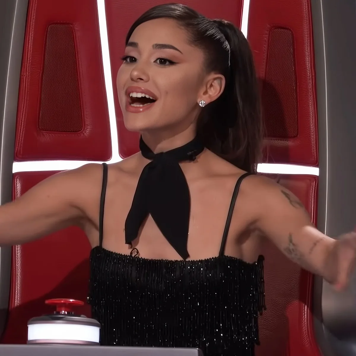 A woman in a black dress with a bow, sitting in a red chair, gesturing with her arms outstretched.