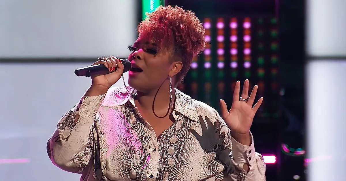 A woman with red hair and a snake print shirt sings into a microphone on stage.