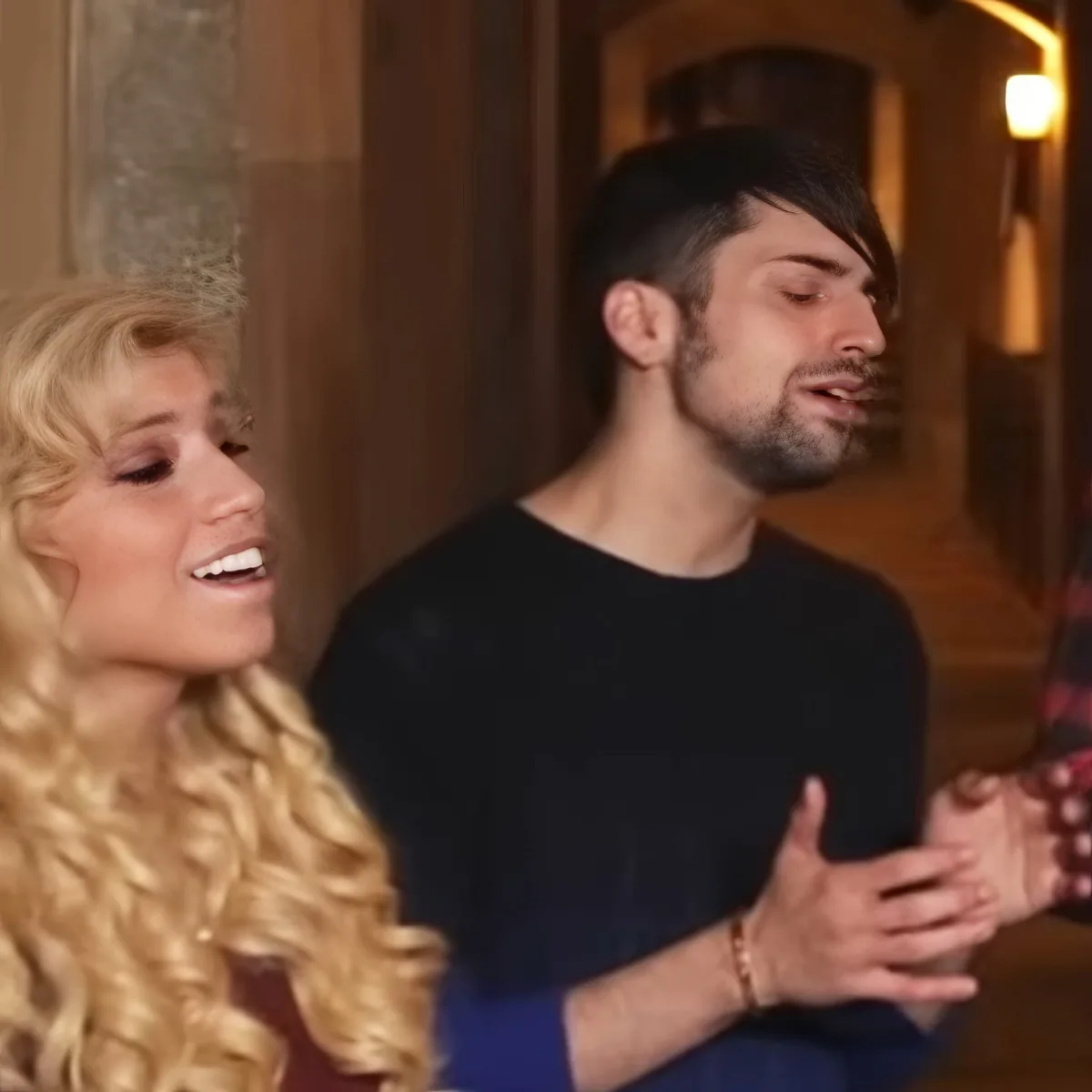 A man in a black shirt and a woman with long blonde hair are holding hands, appearing to be in a hallway.