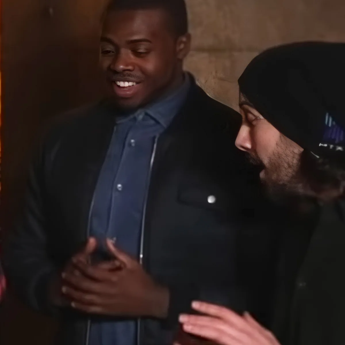 A man in a blue shirt and black jacket gestures while talking to another person wearing a beanie.