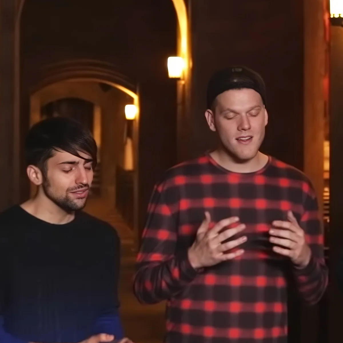 A man in a red plaid shirt gestures with his hands while another man in a black shirt looks on in a dimly lit hallway.