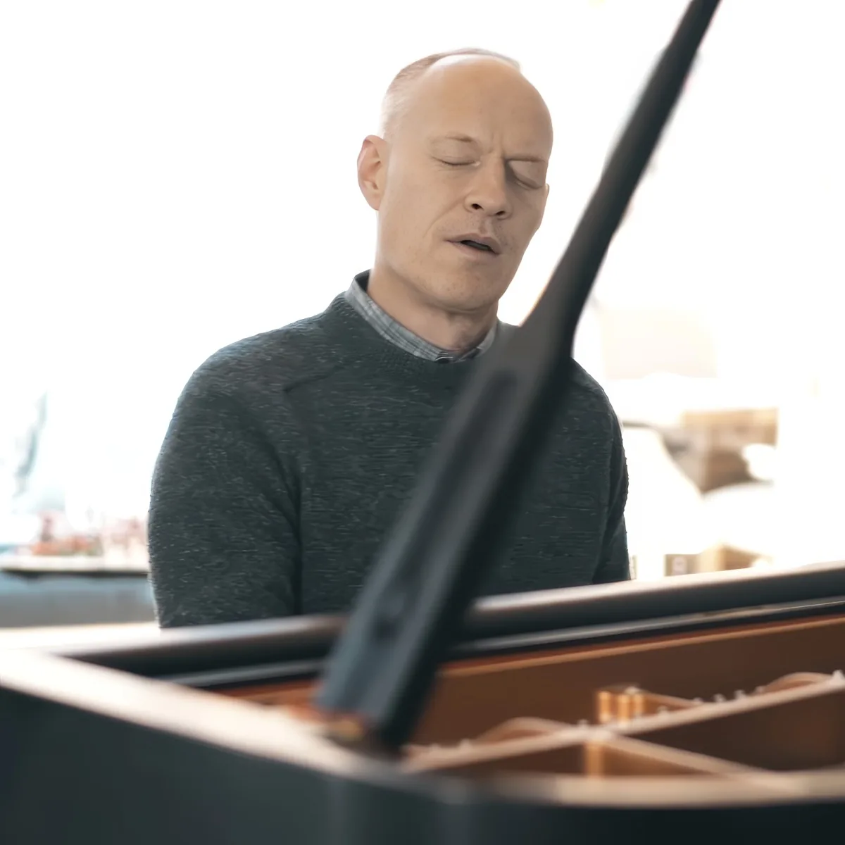A man in a dark sweater stands behind a piano, eyes closed, appearing to sing or concentrate.