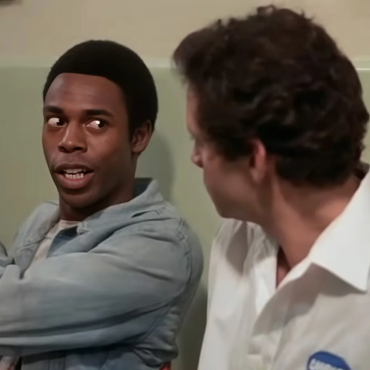 A man in a light blue shirt sits with arms crossed, looking to his right, while another man in a white shirt speaks to him.