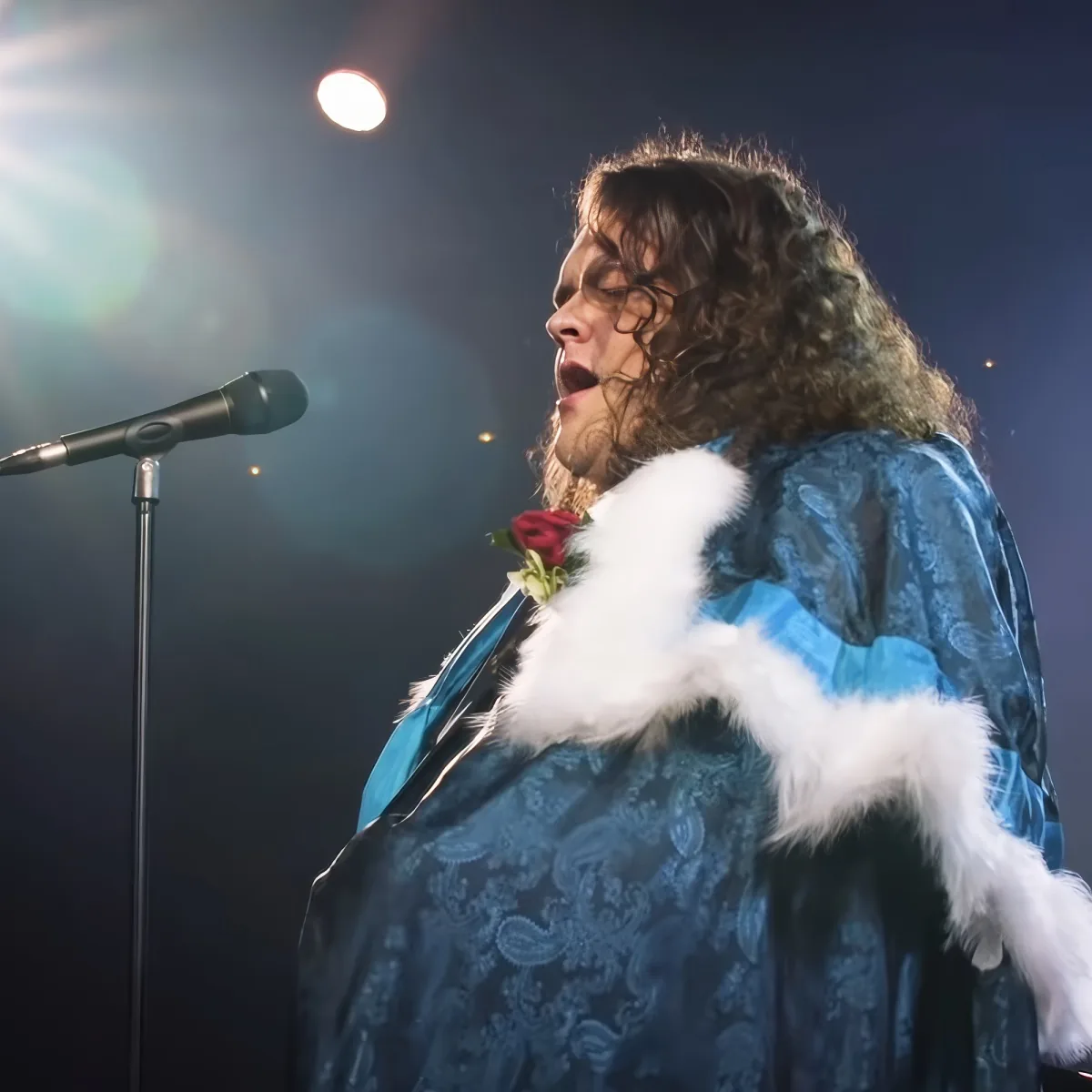 A person in a blue, patterned robe with a white fur collar sings into a microphone on stage.