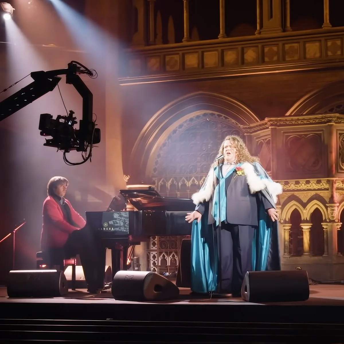 A person in a blue robe sings on stage with a pianist, surrounded by ornate architecture and stage equipment.