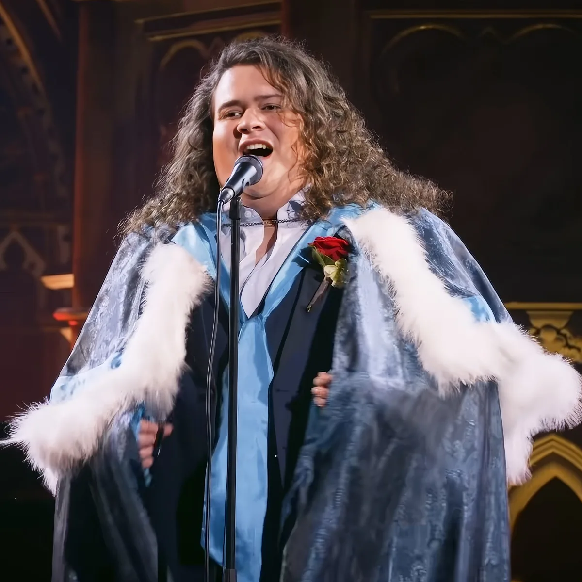 A person in a blue robe with white fur trim sings into a microphone, adorned with a red rose.
