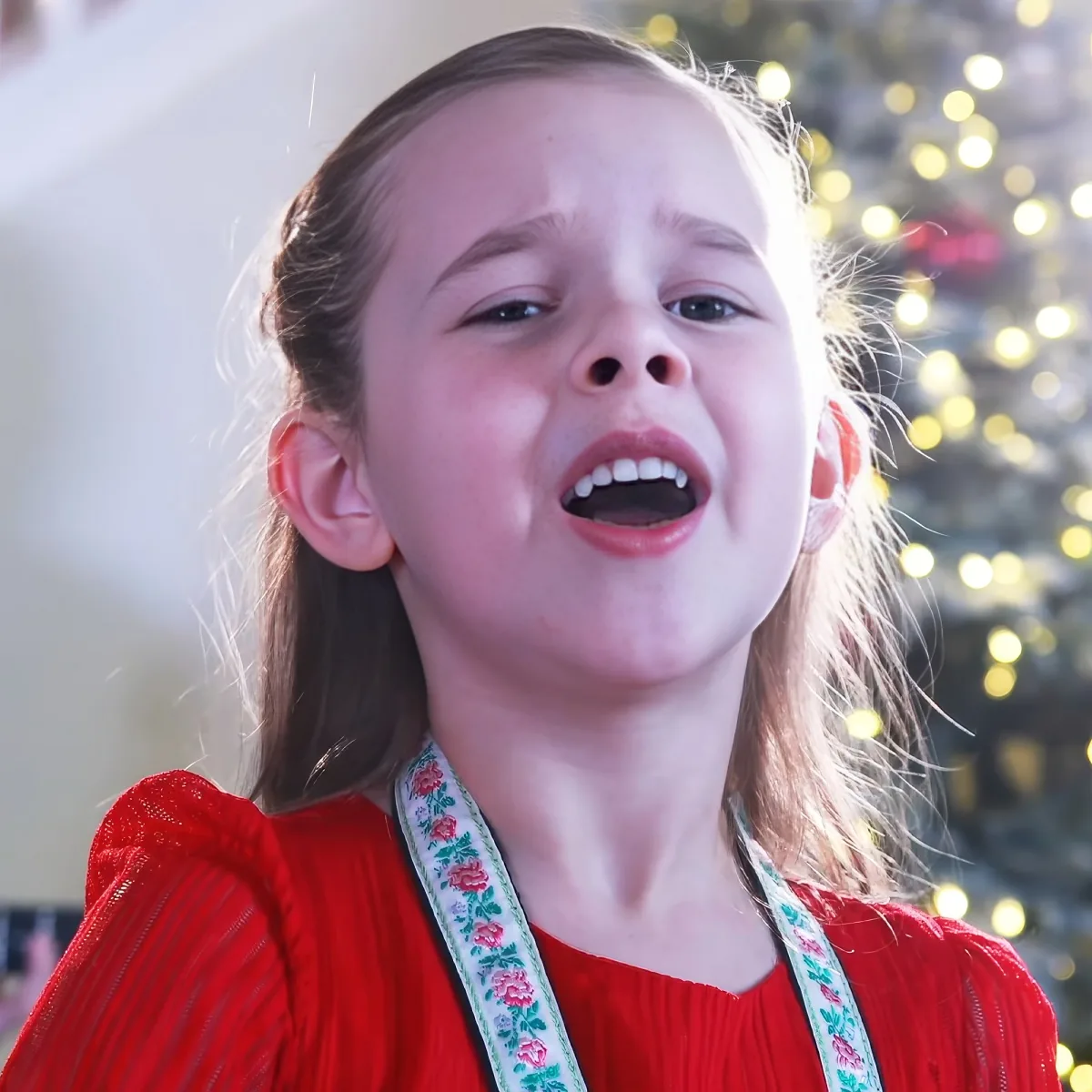 A young girl in a red dress sings with a Christmas tree in the background.
