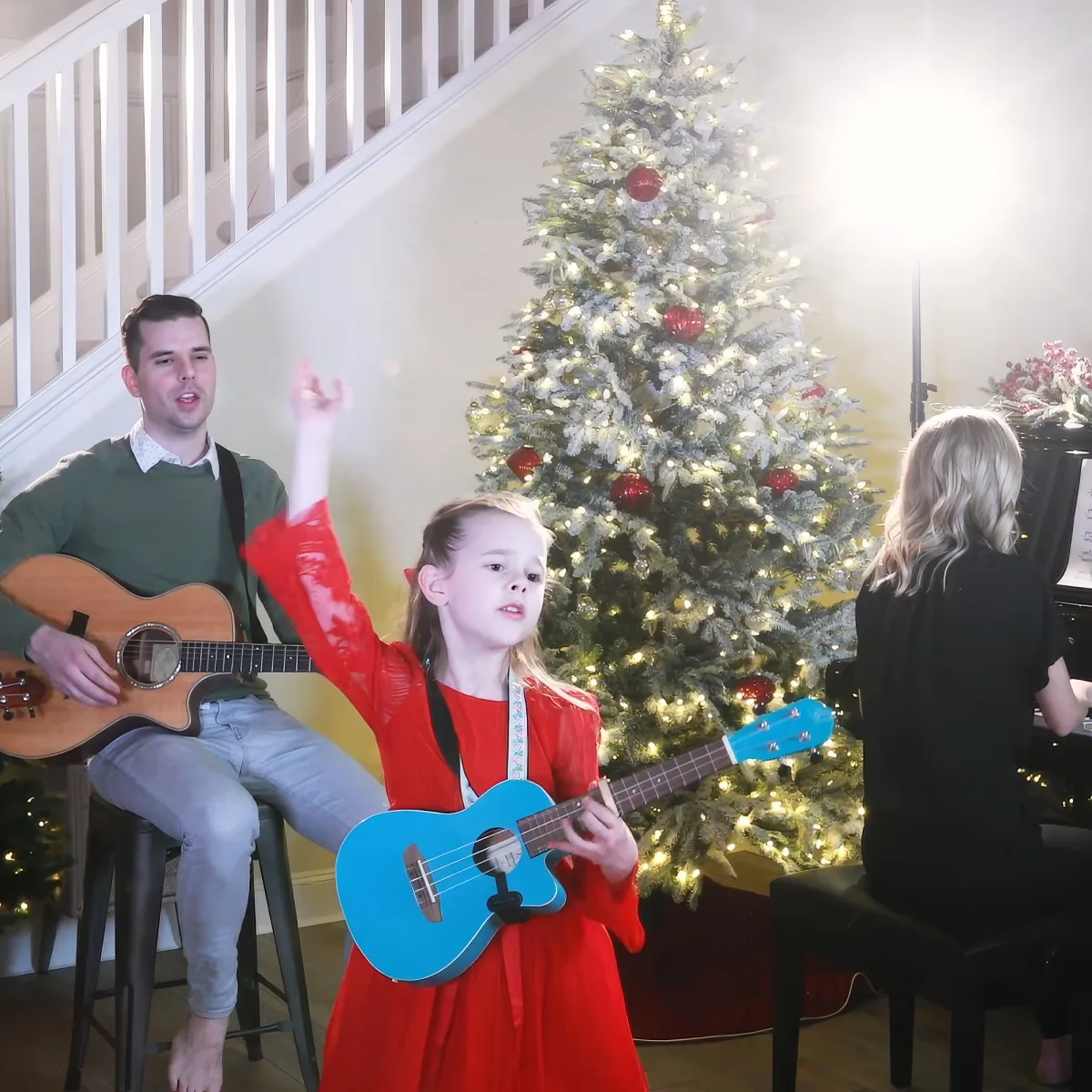 A young girl in red plays a blue ukulele in front of a Christmas tree, with a man playing guitar and a woman at a piano.