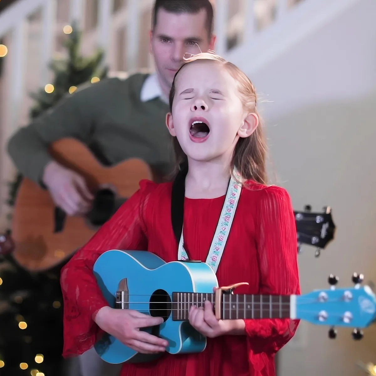 A young girl in red plays a blue ukulele while singing, with a man playing guitar in the background.