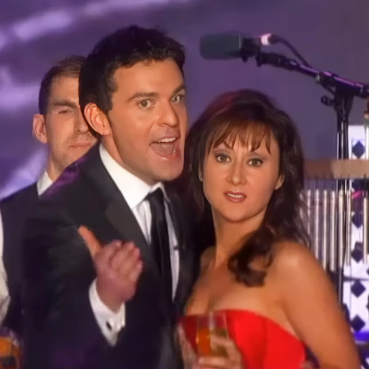 A man in a suit and tie gestures while standing next to a woman in a red dress at what appears to be a formal event.