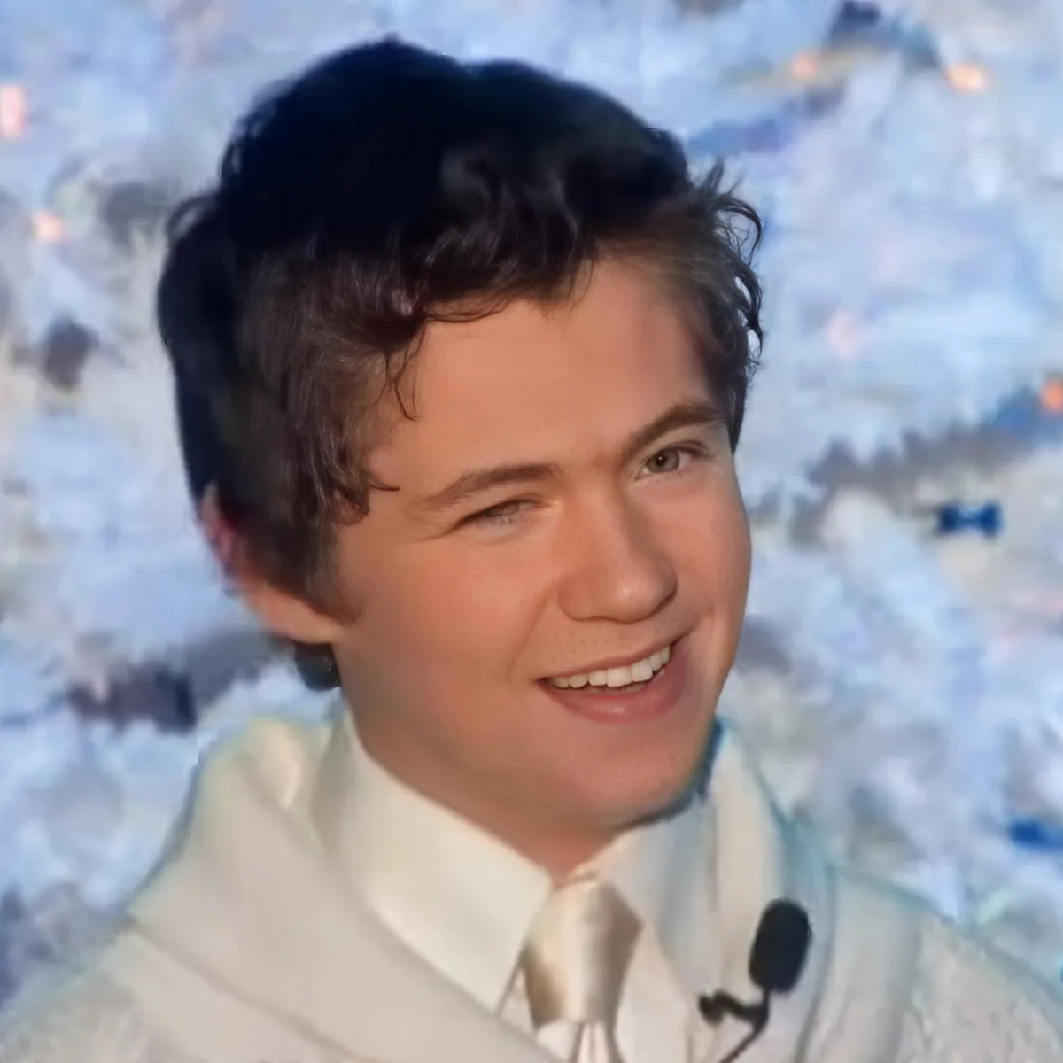 A smiling person in a white suit and tie, with a microphone, against a snowy background.