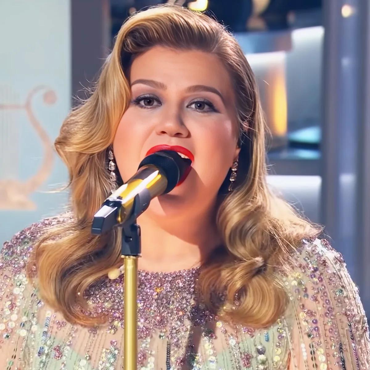 A woman with long, wavy blonde hair sings into a microphone, wearing a sparkly, beaded dress.