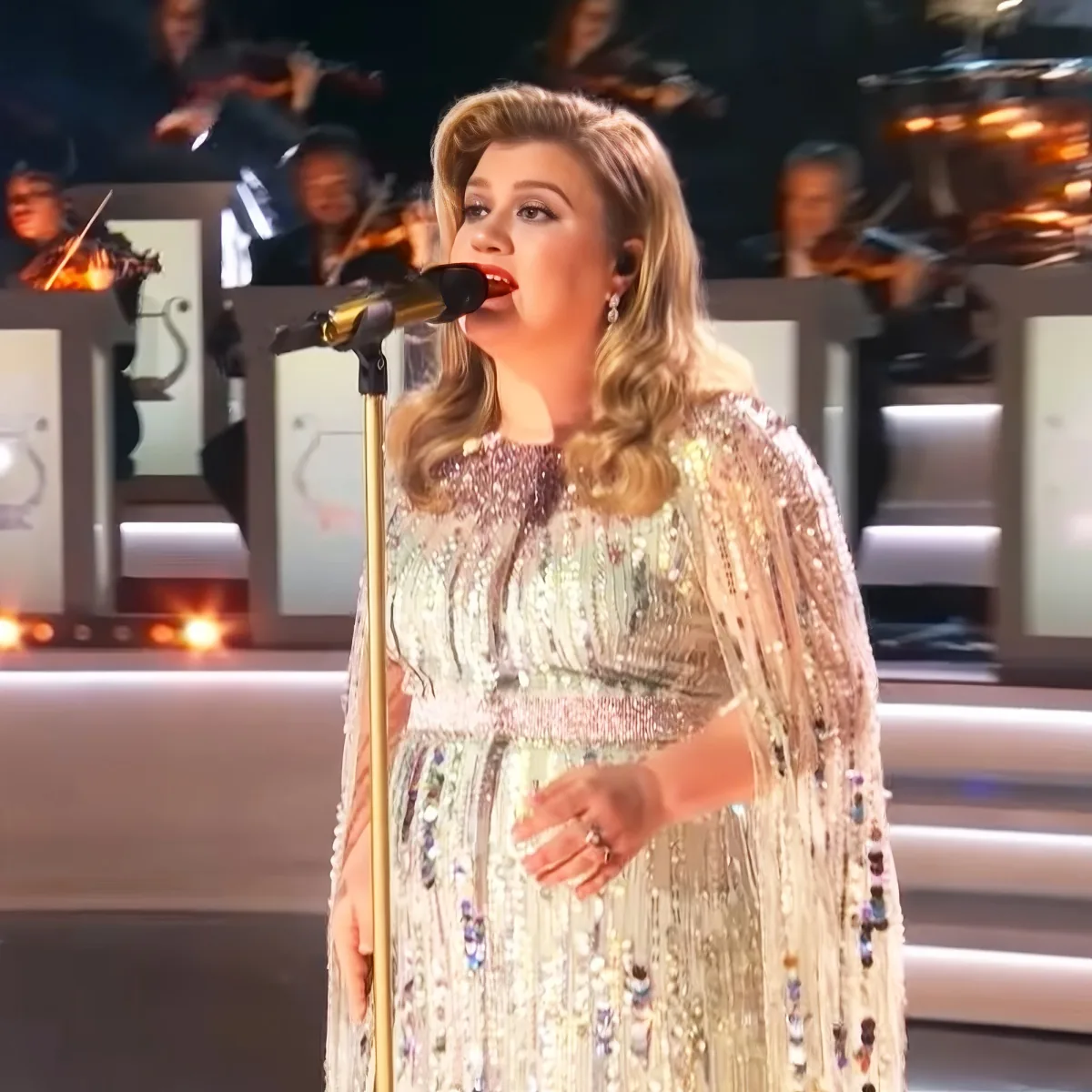 A woman in a sparkling, beaded dress sings into a microphone, with an orchestra in the background.
