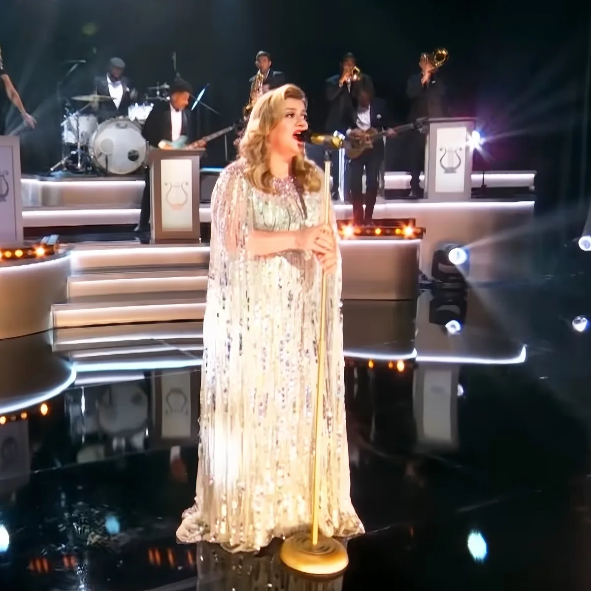 A woman in a sparkly dress sings on stage with a band behind her.