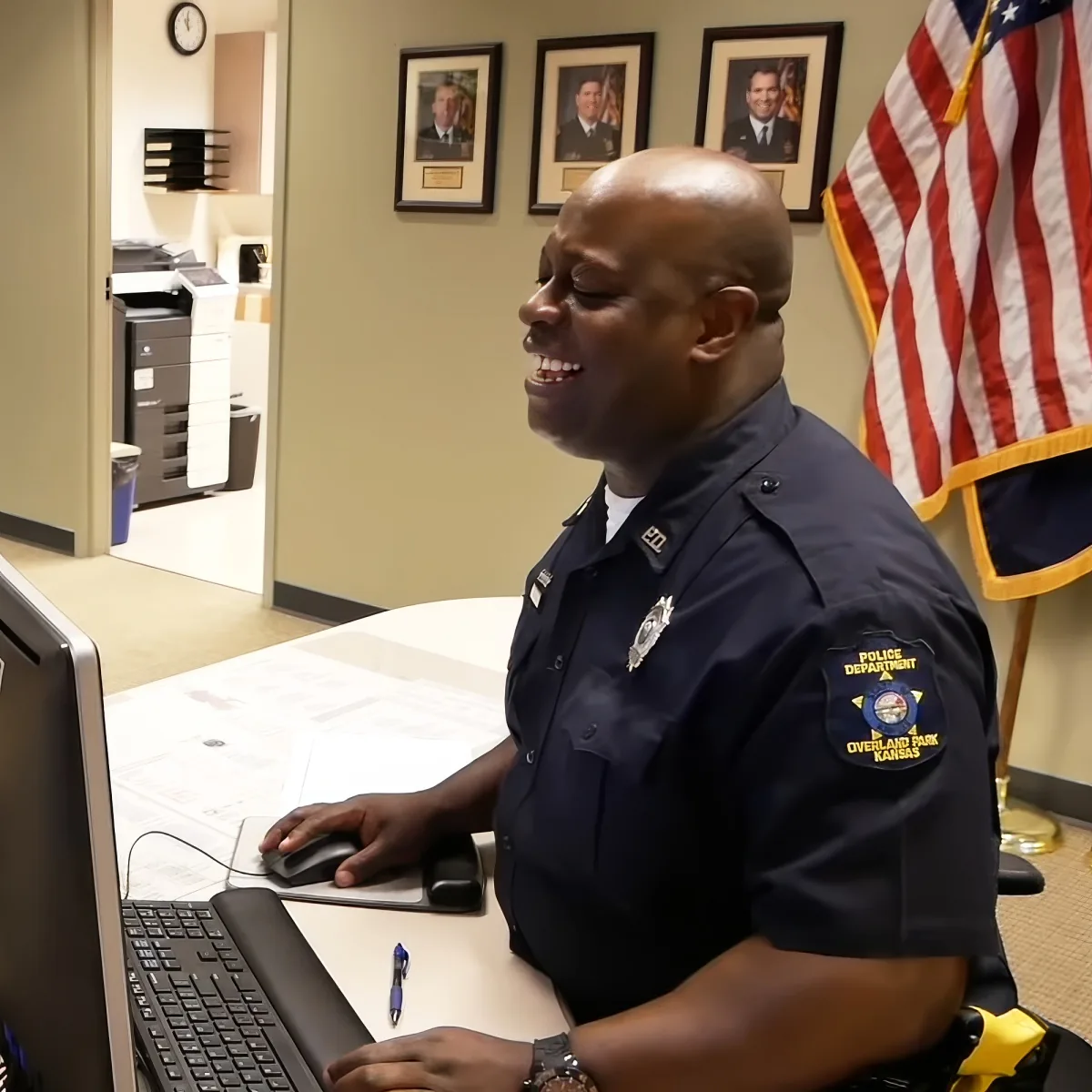 A police officer in Overland Park, Kansas, smiles while working at a desk with a computer and an American flag nearby.