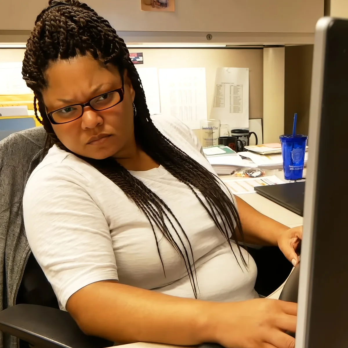 A woman with braided hair and glasses is seated at a desk, working on a computer.