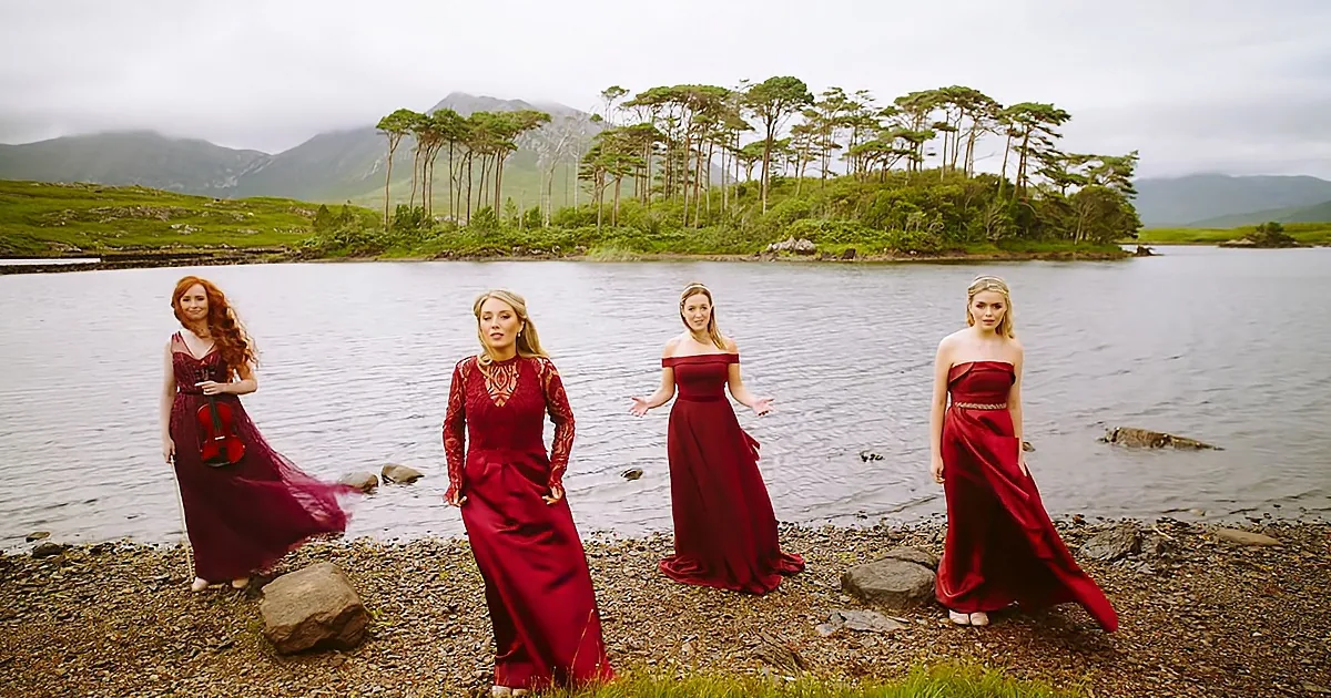 Four women in red dresses stand by a lake, one holding a violin, against a backdrop of trees and mountains.