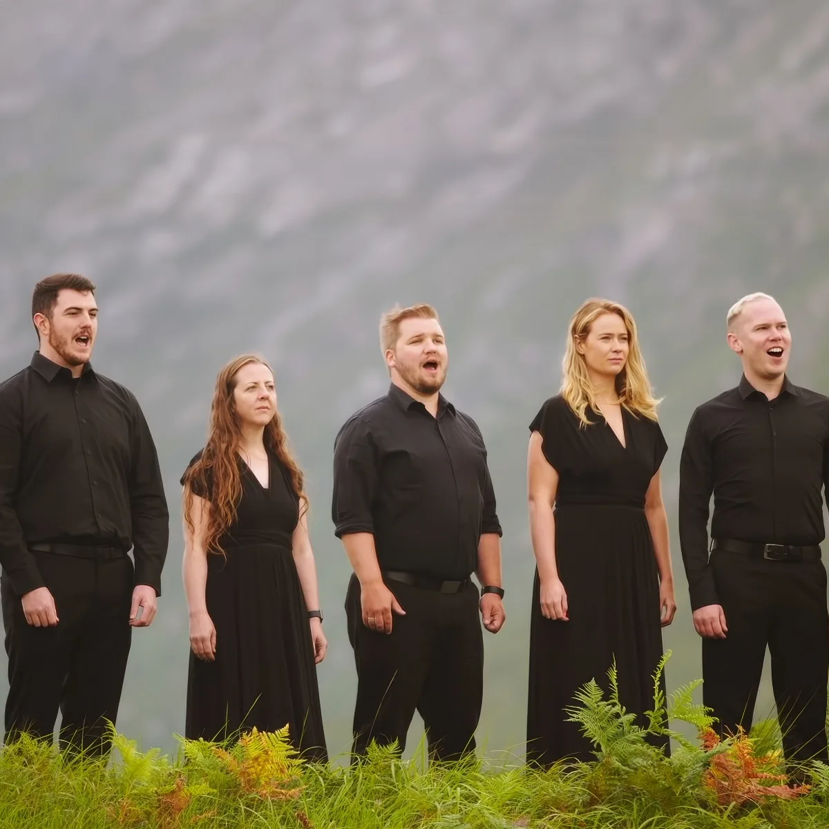 A group of five people in black attire stands outdoors, singing with a mountainous backdrop.